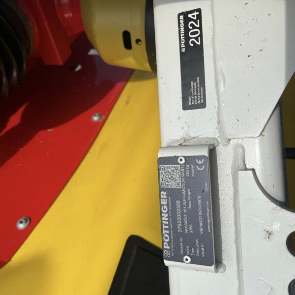 Close-up of a gray machine component with a black label showing the number 2024 and the Pottinger branding, against a yellow and red surface in the background (industrial equipment).