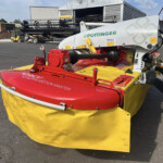 Front section of a Pöttinger NovaCat 301 Alpha Motion Master mower in red and yellow, parked in an equipment yard at a farm/industrial site.