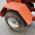 Orange metal machinery frame with a worn rubber tire and rust, resting on a textured concrete surface.