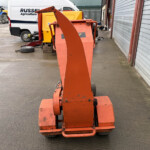 Front view of an orange mobile lifting platform in a service yard, with a white van and blue tractors in the background.