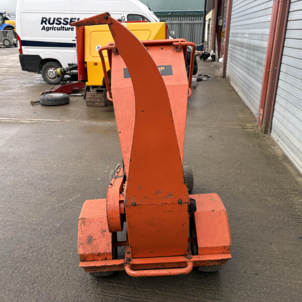 Front view of an orange mobile lifting platform in a service yard, with a white van and blue tractors in the background.