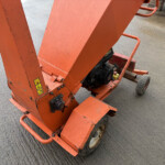 Orange industrial lifting machine on a wheeled base, viewed from a low angle on a textured concrete surface with warning stickers visible on the frame.