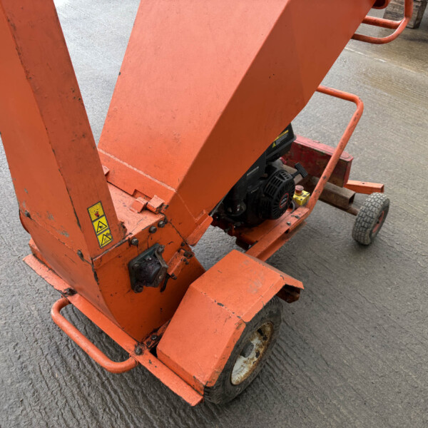 Orange industrial lifting machine on a wheeled base, viewed from a low angle on a textured concrete surface with warning stickers visible on the frame.