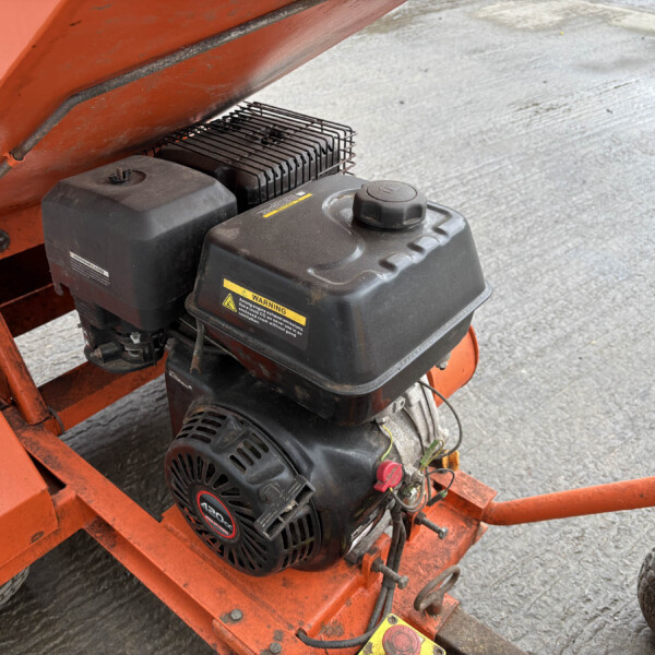 Gas-powered engine mounted on orange industrial equipment, with warning labels and a metal cooling fin cover visible on a dusty concrete surface