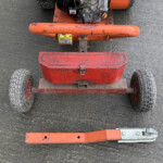 Top-down view of a red wheeled cart with an orange handlebar, engine mounted on top, on a dirt surface.
