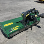 Green industrial mower attachment on a paved surface, viewed from the side with warning decals and hydraulic lines visible.