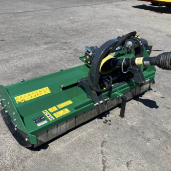 Green industrial mower attachment on a paved surface, viewed from the side with warning decals and hydraulic lines visible.