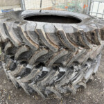 Stack of large muddy tractor tires piled in an outdoor industrial yard near fencing and containers.