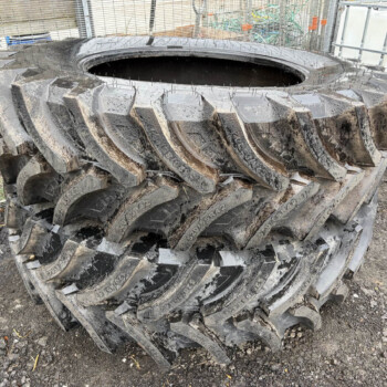 Stack of large muddy tractor tires piled in an outdoor industrial yard near fencing and containers.