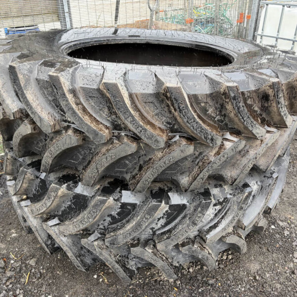 Stack of large muddy tractor tires piled in an outdoor industrial yard near fencing and containers.
