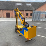 Yellow and blue hydraulic construction machine parked in an empty parking lot in front of a red brick building with a tall metal fence behind it.