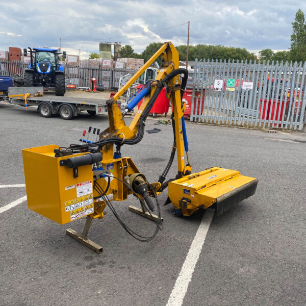 Yellow hydraulic construction machine in a parking lot yard with pallets and a blue pickup on a trailer in the background behind a fence.