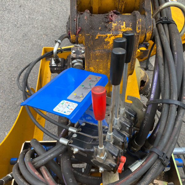 Yellow hydraulic construction machine with a blue control panel, red levers, and many black hoses on a worn rusted frame.