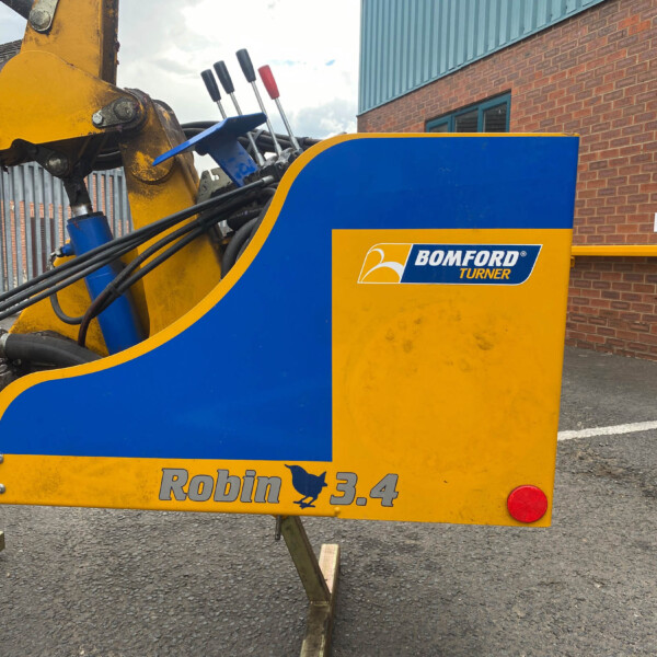 Close-up of a yellow and blue construction machine with hydraulic hoses, displayed near a brick building with signage (Honda, Visitor Parking).