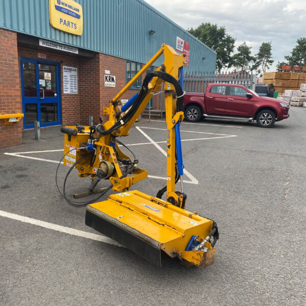 Yellow hydraulic machine in a parking lot outside a brick building with blue trim and a New Holland Agriculture Parts sign, a red pickup in the background and stacked lumber nearby.