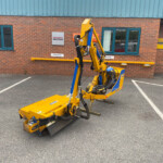 Yellow hydraulic construction machine with exposed hoses parked in a lot in front of a brick building with a blue door and Honda sign behind it.