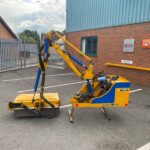 Yellow and blue construction vehicle parked in a lot beside a brick building with Kubota signage and a metal gate in the background.
