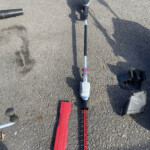 Overhead view of a red hedge trimmer with a long blade lying on gravel, with a gray battery-powered tool nearby and a black case beside it.