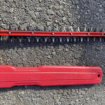Red tool attachment with a row of sharp triangular teeth lying on gravelly ground on a metal frame