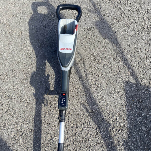 Top-down view of a handheld metal detector resting on rough pavement, showing its black handle and gray shaft with a control panel near the bottom edge of the image.