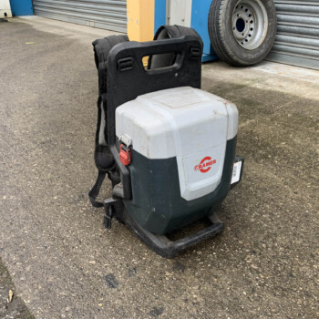 Portable battery pack attached to a black backpack frame on pavement, with a gray hard case and red latch; storefront with a tire in the background.