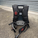 Back view of a black backpack with padded shoulder straps, resting on the ground in front of a gray metal shutter; red logo visible on the left strap.