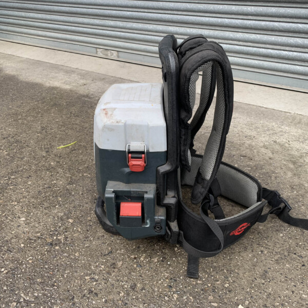 Backpack-style device with a gray-green body and red latch, resting on a rough pavement beside a closed metal shutter; multiple black straps drape over it.