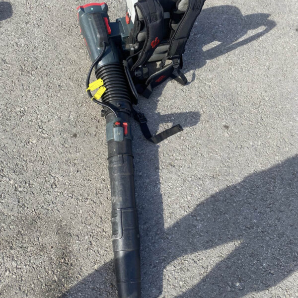 Leaf blower with a long nozzle and a carrying backpack resting on gravel.