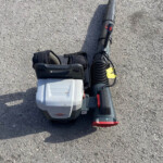 Cordless leaf blower lying on a rough gravel surface with its battery pack attached and a flexible tube trailing alongside.