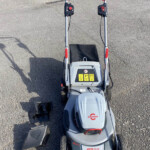 Overhead view of a gray ride-on lawn mower on gravel, showing dual handlebars, red control levers, and warning labels on the console.