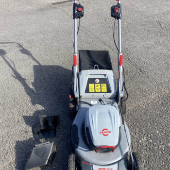 Overhead view of a gray ride-on lawn mower on gravel, showing dual handlebars, red control levers, and warning labels on the console.
