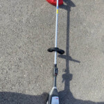 Top-down view of a cordless string trimmer with a red guard resting on rough asphalt driveway surface