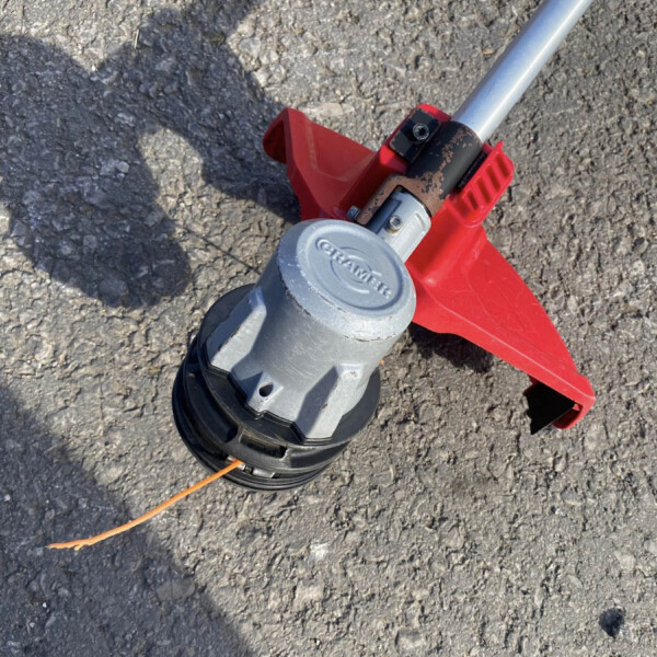 Close-up of a red and gray string trimmer lying on rough asphalt, with an orange cord visible next to the head.