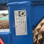 Close-up of a blue metal beam with a silver label attached, showing technical information; a red taillight edge appears in the lower left corner.