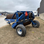 Blue agricultural roller implement with spiral drums and large tires, parked in a yard beside a metal warehouse building.