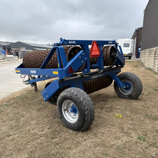 Blue agricultural roller implement with spiral drums and large tires, parked in a yard beside a metal warehouse building.
