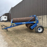 Blue Dal-Bo agricultural roller in the foreground beside a brick retaining wall and gray metal industrial building; a white caravan and a blue truck are parked in the distance.