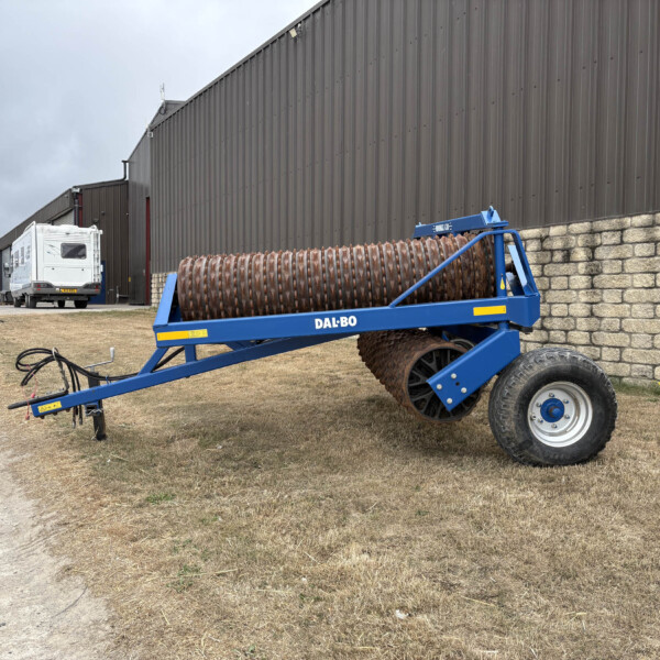 Blue Dal-Bo agricultural roller in the foreground beside a brick retaining wall and gray metal industrial building; a white caravan and a blue truck are parked in the distance.