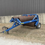 Blue agricultural soil roller attachment (Dal-Bo Minimax 630) with spiral-ridged steel rollers, parked beside a stone wall and metal siding building.