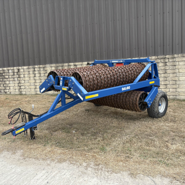 Blue agricultural soil roller attachment (Dal-Bo Minimax 630) with spiral-ridged steel rollers, parked beside a stone wall and metal siding building.