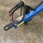 Close-up of blue construction equipment frame with thick black hydraulic hoses and red cords on a dry grassy field, safety labels visible by bolts.