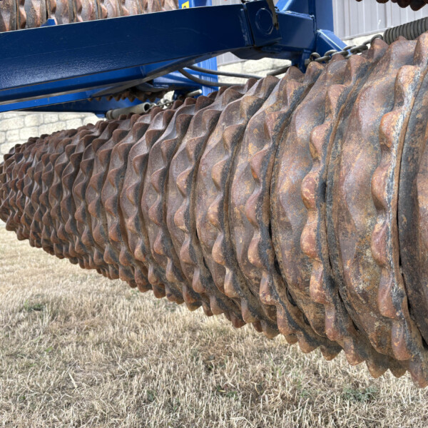 Close-up of a rusted circular roller with wavy blades attached to a blue farm attachment, outdoors.
