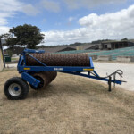 Blue agricultural roller machine with multiple rusted steel drums in a yard, near a brick wall and farm buildings under a cloudy sky.