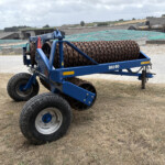 Blue agricultural roller implement on wheels with rusted spiral drums, parked in a farmyard with barns in the background