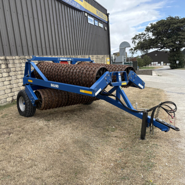 Blue Minimax 630 agricultural roller attachment parked beside a stone wall and grey metal building outside a farm/industrial yard.