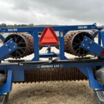 Blue agricultural roller with large rusted spiked rollers and a bright red reflective triangle in the center, outdoors in a yard.
