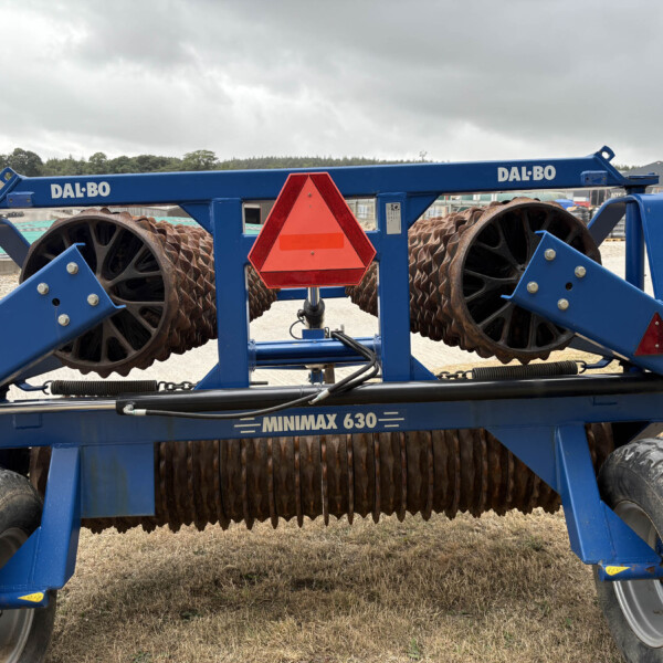 Blue agricultural roller with large rusted spiked rollers and a bright red reflective triangle in the center, outdoors in a yard.