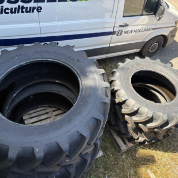 Stacks of large black tractor tires piled beside a white New Holland Agriculture van in a dirt lot