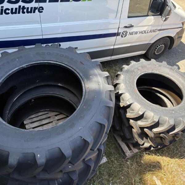 Stacks of large black tractor tires piled beside a white New Holland Agriculture van in a dirt lot