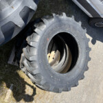 Large rugged tractor or industrial tire with deep treads leaning against a stack of tires on a gravel surface nearby a white vehicle.